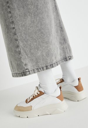 Zapatillas blancas con detalles en beige y marrón, usadas con calcetas blancas hasta el tobillo y vaqueros de pierna ancha de color gris desgastado, vistas desde atrás y de lado.