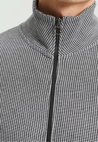 Svart och vit zip-up jacka med ett hundtandsmönster, hög krage, slät textil och en central dragkedja med metalldrag.