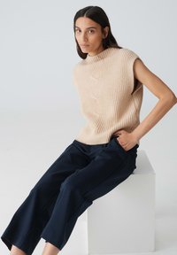 Haut sans manches en tricot beige côtelé avec un col montant, associé à un pantalon cropped navy, posé sur un cube blanc.