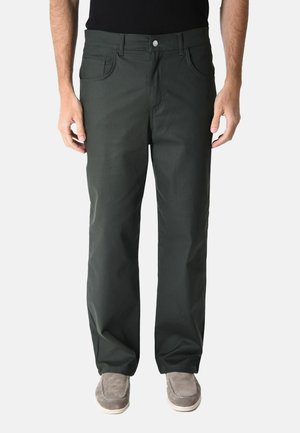 Uomo che indossa pantaloni verde scuro a gamba dritta con tasche frontali e scarpe slip-on in camoscio grigio, in piedi davanti a uno sfondo uniforme.