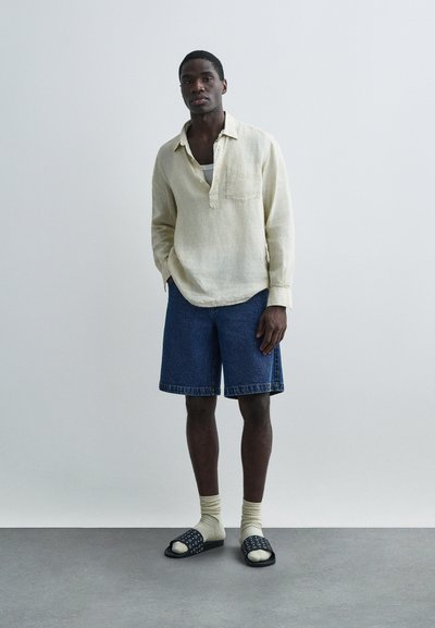 Light beige linen shirt with a chest pocket, paired with dark blue denim shorts. Black slides with white patterns, and cream socks.