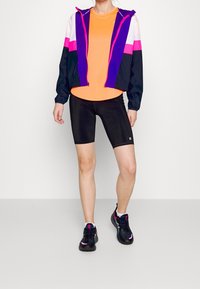 Conjunto deportivo colorido que incluye una chaqueta morada y rosa, camiseta naranja, pantalones cortos de ciclismo negros y zapatillas negras con detalles en rosa.