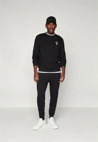 Sweatshirt preta com um colarinho branco, apresentando um pequeno logotipo, combinada com calças de fato pretas e ténis brancos. Design simples e atlético.