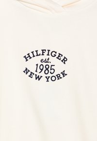 Felpa color crema con logo blu navy che recita "HILFIGER est. 1985 NEW YORK." Tessuto morbido, design casual, senza ulteriori pattern.