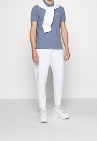 Camisa polo azul con acentos blancos, sudadera blanca colgada sobre los hombros y pantalones de chándal blancos. Llevado con zapatillas blancas.