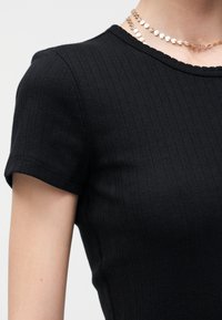 Κοντινό πλάνο ενός ατόμου που φοράει μαύρο ριμπ μπλουζάκι με κοντά μανίκια και χρυσή αλυσίδα με μικρά κρεμαστά κοσμήματα σε σχήμα καρδιάς.