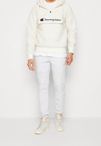 Man i vit fleece Champion-hoodie, ljusgråa sweatpants och krämfärgade Nike högtoppsskor, stående mot en neutral bakgrund.