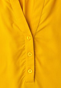 Chemise jaune vif en gros plan montrant trois boutons et une patte cousue sur un tissu lisse avec un col en V.