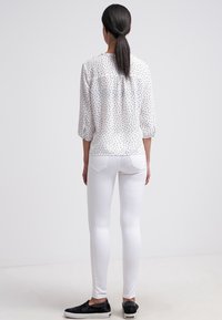 Blouse blanche à pois noirs, manches trois quarts, avec un dos froncé, associée à des leggings blancs et des chaussures noires sans lacets.