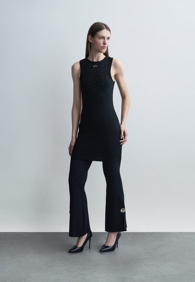 Γυναίκα που στέκεται μπροστά σε γκρίζο τοίχο, φοράει μαύρο αμάνικο τούνικ με μικρές τρουκς πάνω από μαύρο παντελόνι καμπάνα και μαύρα ψηλοτάκουνα.