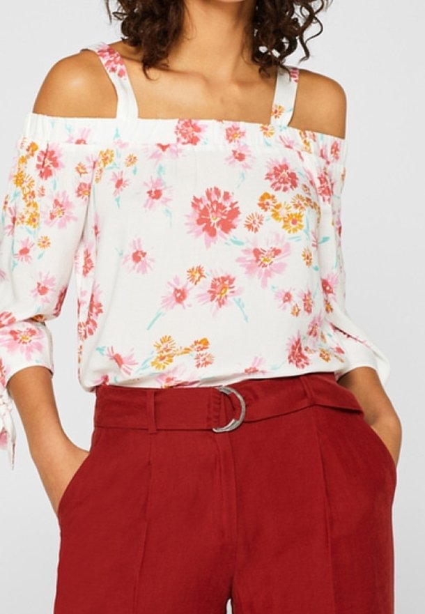 Femme portant un chemisier blanc à épaules dénudées avec des fleurs roses et oranges, associé à un pantalon rouge taille haute avec une ceinture en tissu.