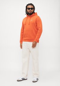 Uomo con capelli e barba intrecciati che indossa occhiali da sole neri, felpa arancione, pantaloni bianchi e sneakers bianche con strisce nere, in piedi su uno sfondo bianco.