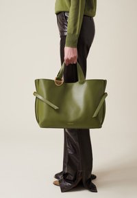 Green leather tote bag with two handles, side straps, and a gold ring accent. Smooth texture and minimalistic design.