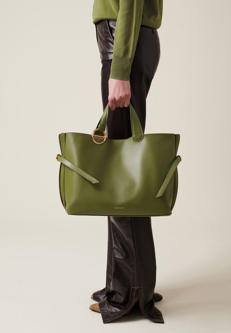 Green leather tote bag with two handles, side straps, and a gold ring accent. Smooth texture and minimalistic design.