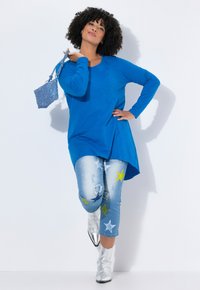 Blaues Langarmshirt mit abgerundetem Saum, kombiniert mit verwaschenen hellblauen Jeans mit Sternmustern in Gelb und Weiß sowie silbernen Knöchelschuhen.