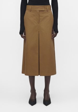 Spódnica do kolan w kolorze koniakowym z przednią centralną plisą, noszona z czarną bluzką z długim rękawem, cienkimi czarnymi rajstopami i czarnymi szpilkami z ostrym noskiem.