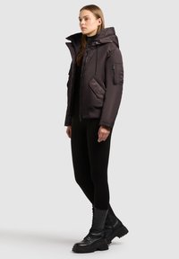 Dunkellila gepolsterte Jacke mit hohem Kragen, mehreren Taschen und verstellbaren Kordelzügen, kombiniert mit schwarzen enganliegenden Leggings und Kampfstiefeln.