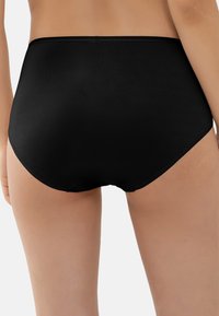 Frau, die schwarze, nahtlose Unterwäsche mit hoher Taille trägt, steht mit dem Rücken zur Kamera vor einem schlichten weißen Hintergrund.