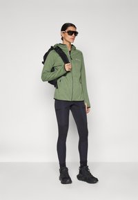 Casaco verde com fecho éclair e capuz, leggings pretas e botas de caminhada pretas. Os acessórios incluem uma mochila preta e óculos de sol oversized.