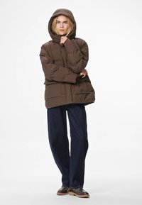 Chaqueta acolchada marrón con capucha, de diseño ancho y grandes bolsillos frontales, combinada con pantalones de mezclilla oscuros y zapatos marrones.