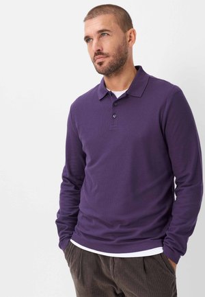 Man met kort haar en baard, draagt een paarse poloshirt met lange mouwen en een bruine corduroy broek, staand met handen in de zakken.