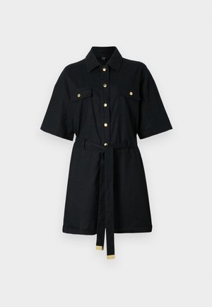 Robe noire à manches courtes avec un devant à boutons, deux poches poitrine et une ceinture en tissu. Portée avec des chaussures noires et un sac à main de couleur claire.