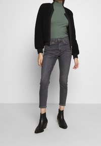 Černá texturová bomber bunda, lesně zelený přiléhavý rolák, šedé skinny džíny a černé kotníkové boty s blokovými podpatky, postávající na neutrálním pozadí.