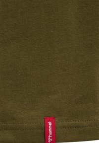 Olijfgroene stof met horizontale stiksels en een klein verticaal rood label met de tekst "hummel" nabij de onderrand.