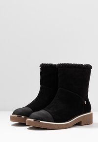 Svarta ankelboots med fuzzy foder, texturerade bruna sålar, förstärkta tåskydd och en liten metallisk logotyp på sidan.