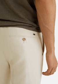 Beige linnen broeken met een achterzak met een knoopsluiting. Subtiele naaddetails en een klein logo-tag nabij de zak.
