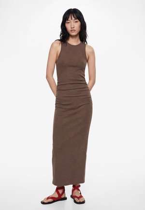 Femme debout, les mains derrière le dos, portant une robe longue marron sans manches et des sandales plates rouges à lanières sur fond blanc.