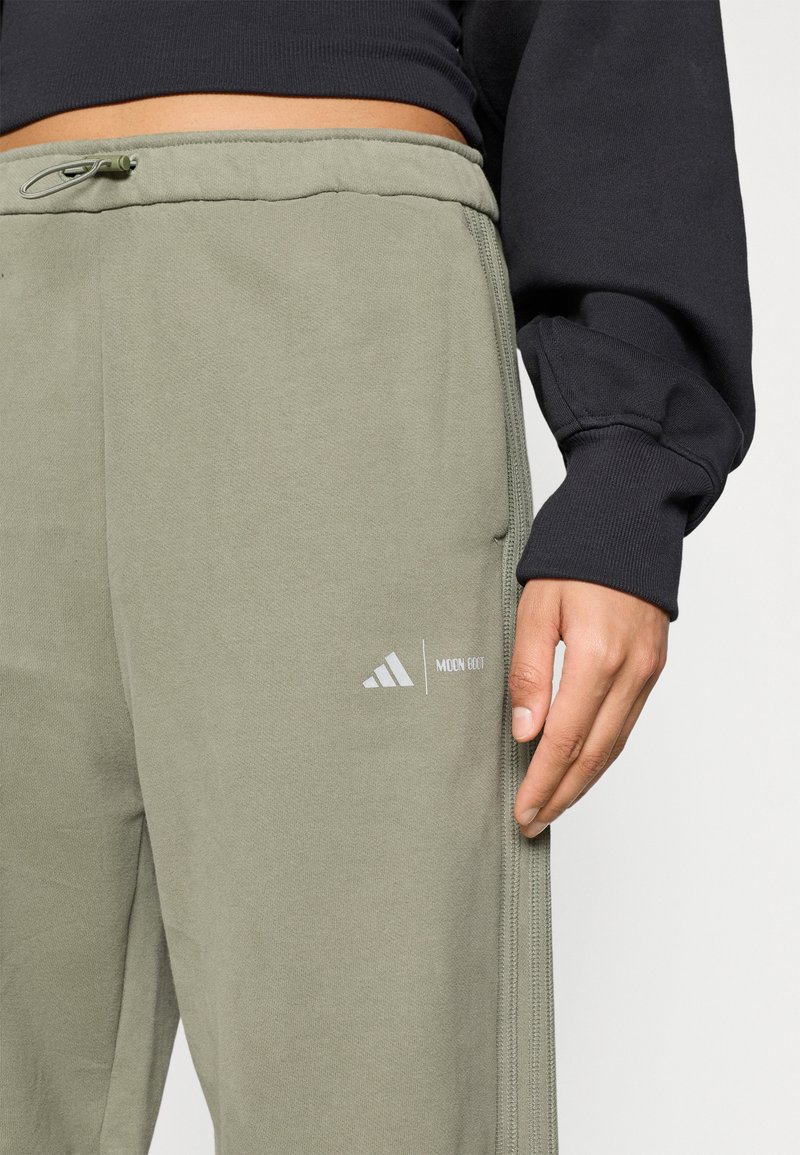 Close-up van een persoon in olijfgroene sweatpants met Adidas en Moon Boot logo's, gecombineerd met een zwart shirt met lange mouwen.