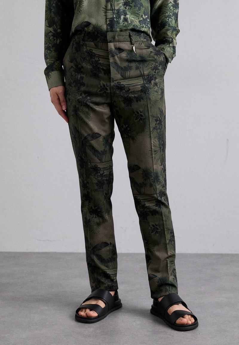 Green patterned trousers featuring botanical designs, slim fit, with a smooth texture, paired with black slide sandals.