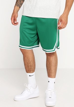 Homme portant un short de sport vert avec bordure blanche, chaussettes Nike blanches et baskets blanches, debout sur un fond uni.