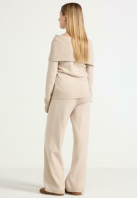 Frau mit langen blonden Haaren, die einen beigen, gestrickten, schulterfreien Pullover und passende, weit geschnittene Hosen trägt, steht vor einem weißen Hintergrund.