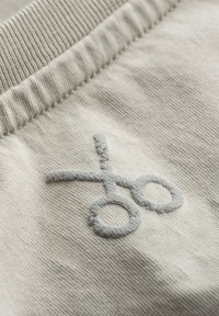 Lichtbeige stof met ribbelstructuur en een grijs geborduurd scharenlogo. De stitching is zacht en pluizig tegen het oppervlak.