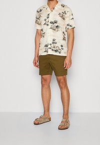 Korte mouw beige shirt met palmbomenprint, olijfgroene shorts en licht beige sandalen met gespen. Glad materiaal en casual pasvorm.