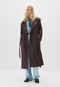 Trench coat lungo in finta pelle marrone con design doppiopetto, ampie revers e vita con cintura, abbinato a jeans blu chiaro.