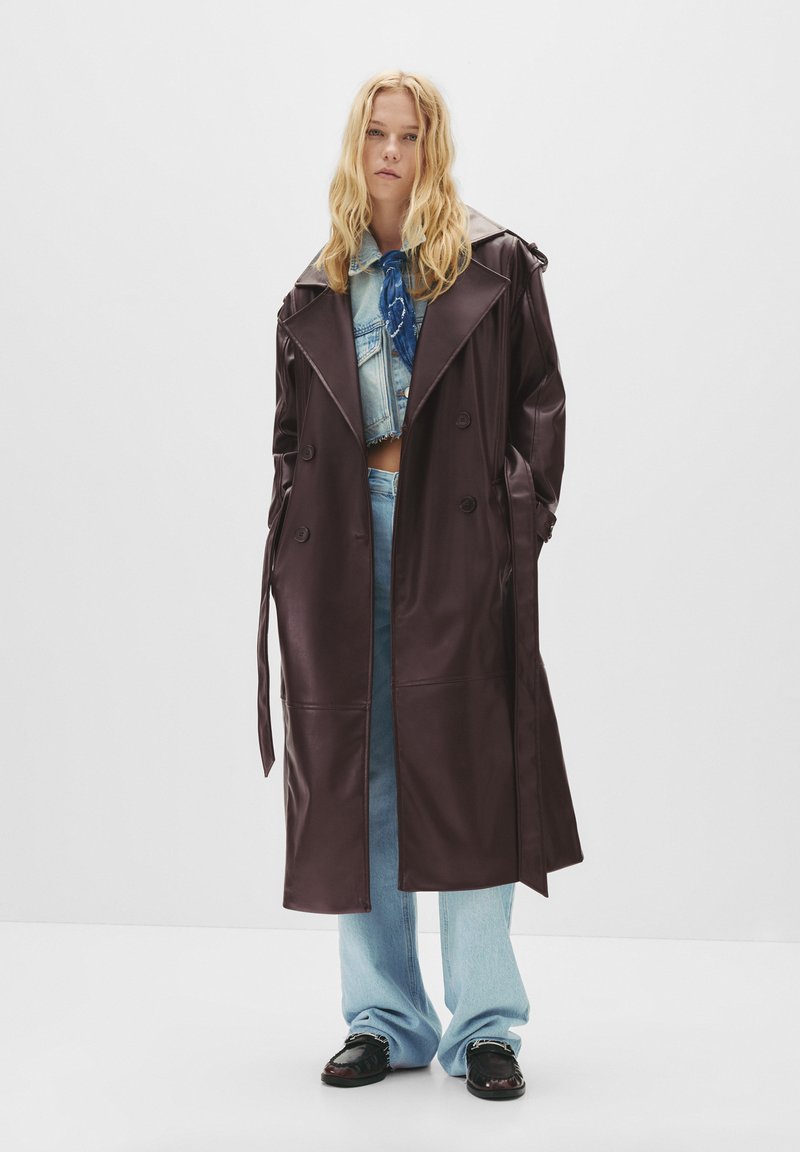 Trench coat lungo in finta pelle marrone con design doppiopetto, ampie revers e vita con cintura, abbinato a jeans blu chiaro.