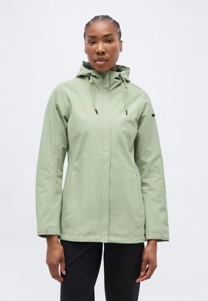 Nő világoszöld kapucnis dzsekiben, elülső zsebekkel és fekete nadrágban, sima háttér előtt áll.