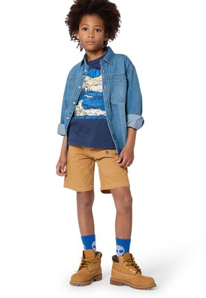 Enfant aux cheveux bouclés portant une chemise en denim par-dessus un t-shirt graphique bleu marine, un short beige, des chaussettes bleues et des bottes beiges à lacets, debout face à l'avant.
