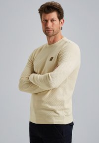 Licht beige textuurtrui met een ronde hals, lange mouwen en een klein logoopdruk op de borst. Draagt bij donkere broek.