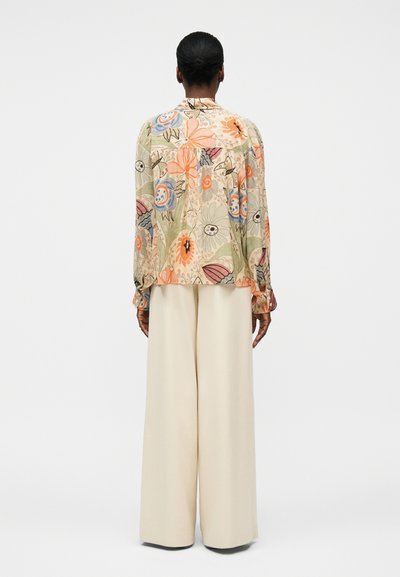 Femme debout de dos, portant un chemisier à manches longues à motifs floraux orange, bleu et vert, associé à un pantalon large beige.