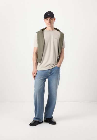 T-shirt beige avec le logo "BOSS", pull vert olive drapé sur les épaules, jeans droit bleu clair, et chaussures noires, sur fond blanc.