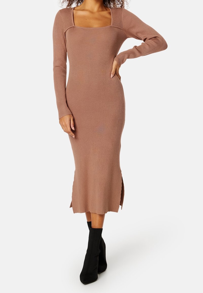 Femme portant une robe midi marron à manches longues côtelées avec des fentes sur les côtés et des bottines noires, debout avec une main sur la hanche.