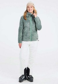 Giacca in fleece verde con colletto alto, chiusura con zip e due tasche laterali; abbinata a pantaloni bianchi e scarponi da sci neri.