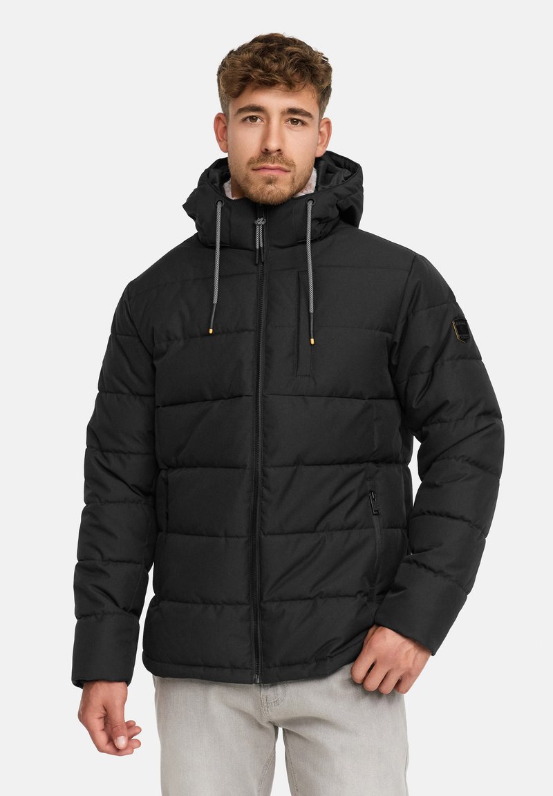 Mann trägt eine schwarze gesteppte Pufferjacke mit Kapuze und Kordelzügen und steht vor einem schlichten weißen Hintergrund.