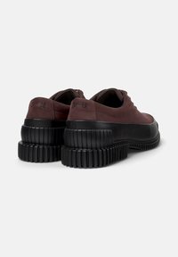 Zapatillas burdeos oscuras con suelas negras gruesas, que presentan una textura suave y el logo de "Camper" en el talón, vistas desde atrás.
