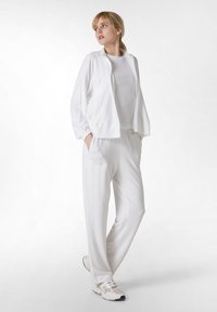 Ensemble de sport blanc comprenant une veste zippée avec des poignets élastiques et un pantalon large assorti, le tout en tissu léger. Baskets avec des accents en maille.