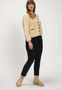 Beige ribbad cardigan med orange fickdetalj, knappfäste fram och uppvikt ärm, tillsammans med svarta tajta byxor och beige snörstövlar.